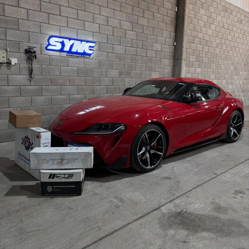 Red Toyota GR Supra A90 in front of the Sync sign, next to boxes of parts